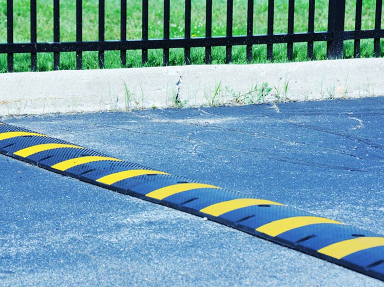 Yellow and black striped speed bump in road next to grassy area behind black fence.